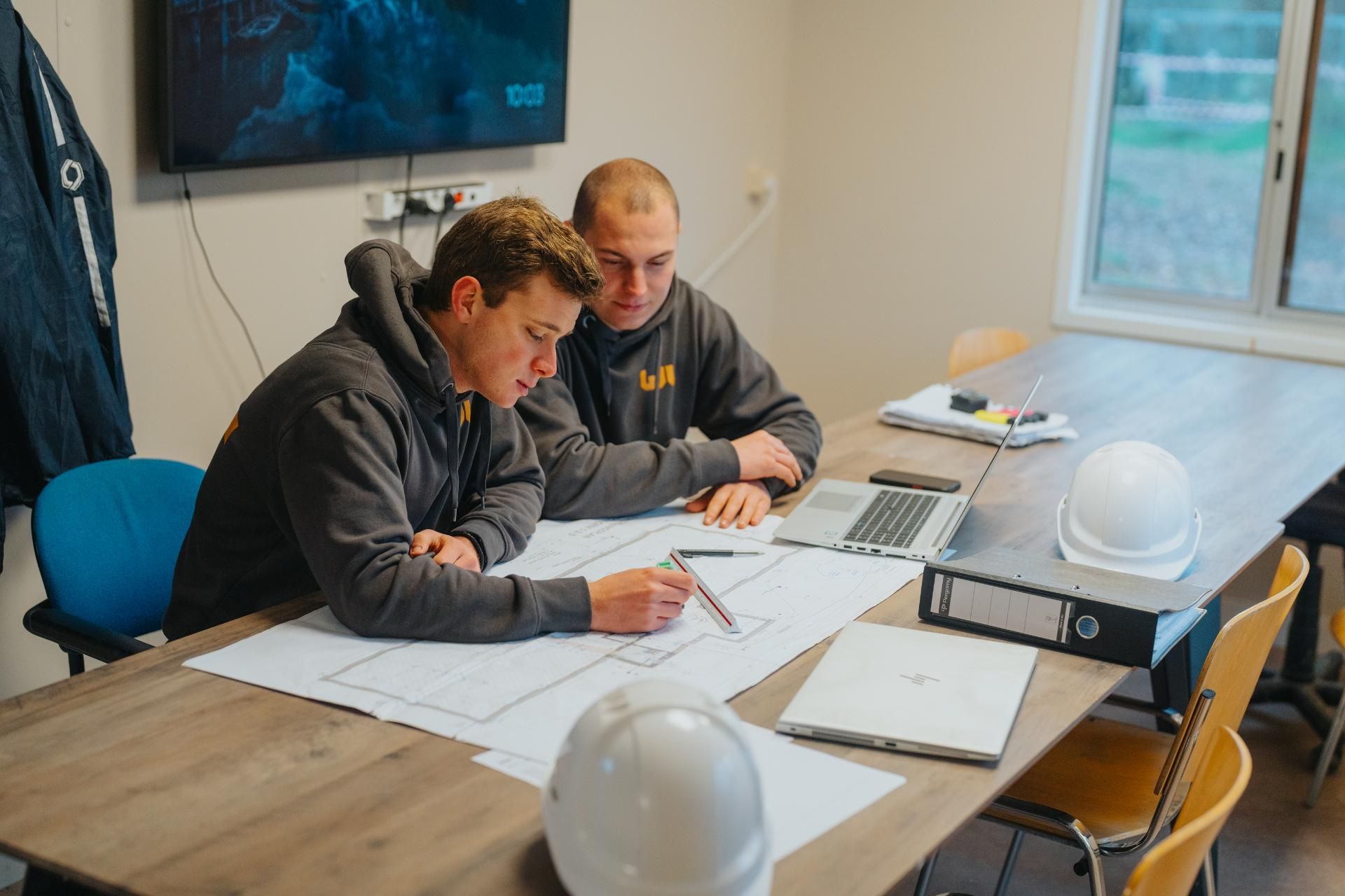 Twee mannen zitten rond een tafel en bekijken architectonische plannen.