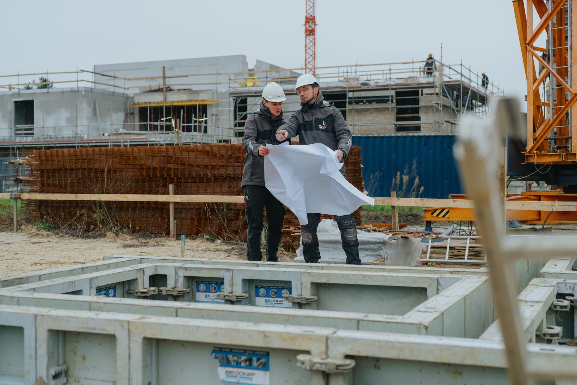 Twee bouwvakkers op een werf die een plan aan het bekijken zijn.