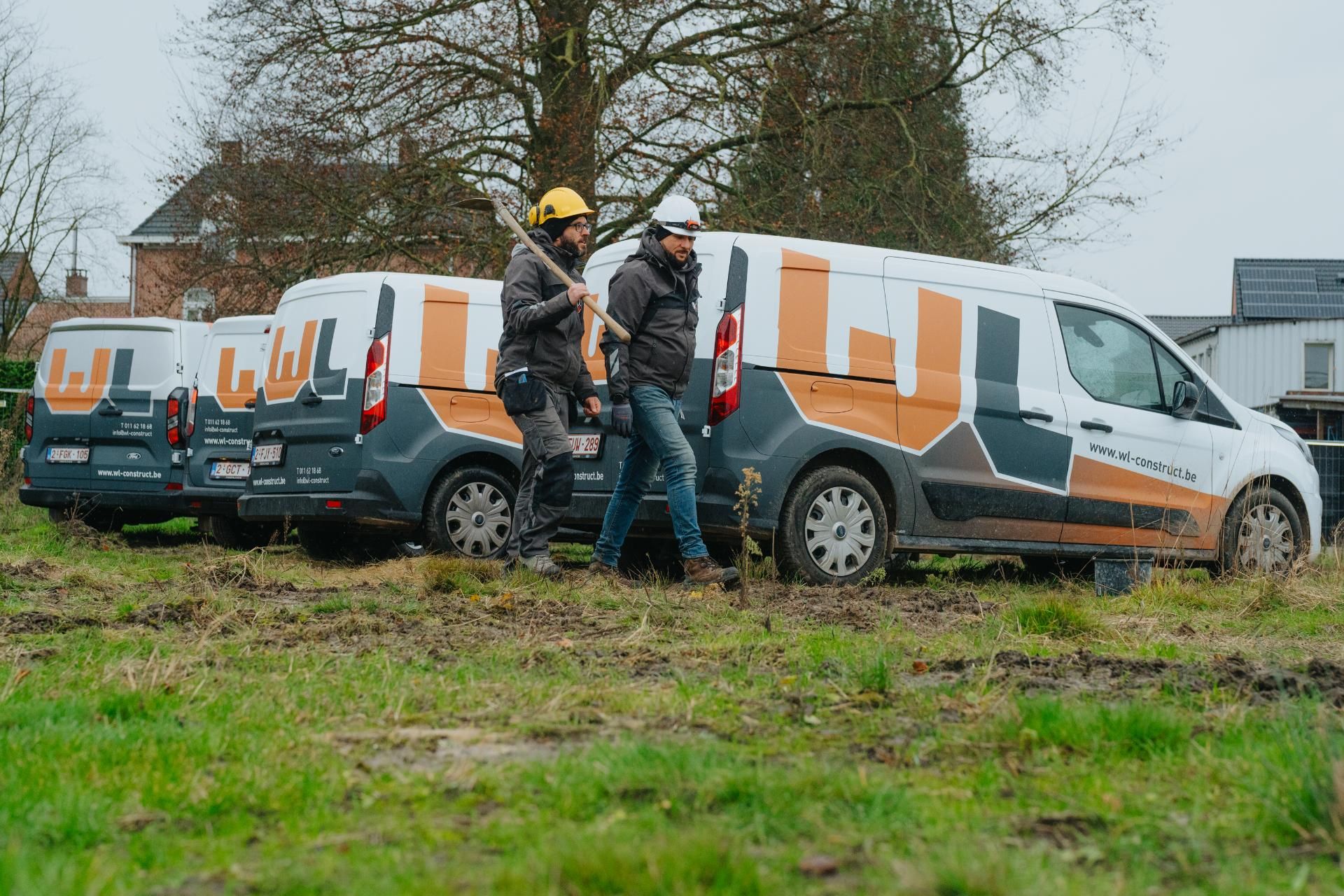 Twee arbeiders in veiligheidskleding lopen over een grasachtig, modderig veld, met gereedschap in de hand. In de achtergrond staan 4 werkbusjes van WL Construct.